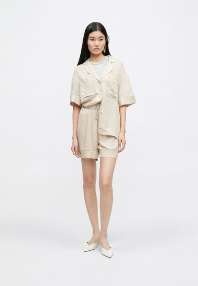 Béžový overal s krátkym rukávom a textúrovaným vzorom, s gombíkovým zapínaním vpredu, s nastaviteľným pásom a dvoma prednými vreckami. Kombinovaný s bielymi slip-on topánkami.