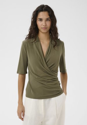 Vrouw met donker krullend haar, gekleed in een olijfgroene top met overslag en ellebooglange mouwen, en witte broek met hoge taille, tegen een effen achtergrond.