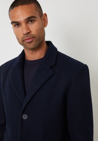 Dunkelblauer Wollmantel mit Reverskragen, einreihigem Knopfverschluss und zwei vorderen Taschen. Getragen über einem strukturierten blauen Pullover.