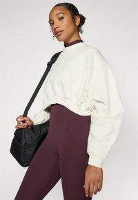 Kremfarget kort sweatshirt med ribbestrikkede mansjetter over dype burgunder høyt liv leggins. Modellen holder en svart veske med stropp.