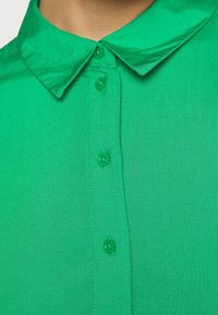 Grüne Hemd mit Kragen, das eine glatte Textur, kurze Ärmel und eine durchgehende Knopfleiste mit passenden grünen Knöpfen aufweist.