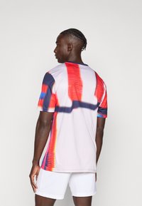 Αθλητική μπλούζα με κοντά μανίκια σε λευκό χρώμα, με αφηρημένα μοτίβα σε κόκκινο, μπλε και μαύρο. Λεία υφή με στρογγυλή λαιμόκοψη.