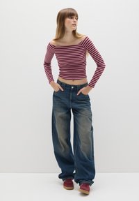 Gestreiftes burgunderfarbenes und cremefarbenes Off-Shoulder-Oberteil, kombiniert mit lockeren blauen Jeans. Das Model steht mit den Händen in den Taschen und trägt rote Sneakers.