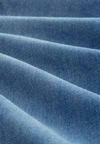 Gros plan sur un tissu denim bleu avec une texture visible et des plis diagonaux créant des ombres douces et des reflets.