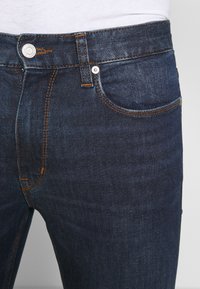 Tmavě modré džíny z denimové látky s oranžovým prošitím, obsahující přední kapsu, kovový knoflík a hladkou strukturu. Žádné viditelné opotřebení.