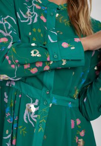 Zöld, gombos elől készült ruha virágos és absztrakt mintákkal rózsaszín, kék és arany színekben. Hosszú ujjakkal és megkötős derékrésszel rendelkezik.