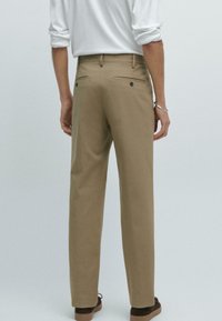 Beige Hose mit geradem Schnitt, ausgestattet mit zwei Gesäßtaschen und subtilen Nähdetails. Getragen mit einem weißen Langarmhemd.