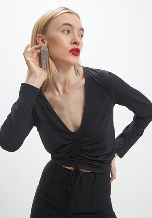 Donna con lunghi capelli biondi, indossa un top nero a V arricciato e pantaloni, mentre aggiusta grandi orecchini a frange argentate, con rossetto rosso brillante.
