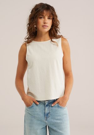 Tânără cu păr creț și castaniu, purtând un top alb fără mâneci și blugi deschiși la culoare, cu mâinile în buzunare, pe un fundal neutru.