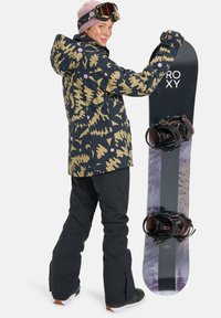Snowboarder in einer gemusterten schwarzen und goldenen Jacke, schwarzen Hosen und einem pinken Hut, der ein schwarzes Snowboard mit lila Akzenten und Bindungen hält.