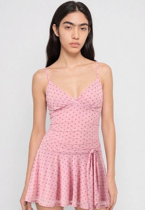 Jeune femme aux longs cheveux foncés portant une robe rose à pois sans manches avec un nœud sur le côté, debout devant un fond clair uni.