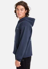 Sudadera azul marino de mezcla de algodón con un bolsillo frontal, capucha con cordón y estampado gráfico en el costado. El modelo posa de perfil.