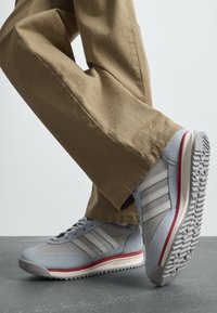 Egy világoskék és szürke Adidas sportcipőt viselő személy, piros és fehér talppal, bézs nadrágban, szürke padlón áll.