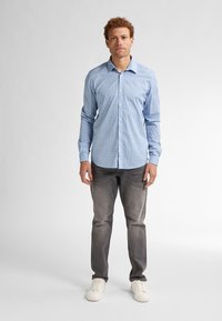 Camicia azzurro chiaro con un motivo a griglia, maniche lunghe, abbinata a jeans grigi e sneaker bianche, in piedi su uno sfondo semplice.