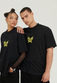 T-shirts noirs avec un graphisme de papillon inspiré des flammes jaunes sur la poitrine ; coupe ample, en coton, avec un modèle portant une manche en mesh.