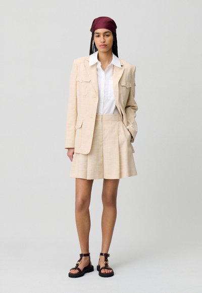 Blazer di lino beige con due tasche frontali, abbinato a pantaloni corti chiari. Camicia bianca sotto. Sandali marroni con cinturini e accenti dorati.