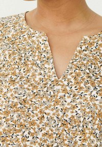 Kwiatowa bluzka z dekoltem w kształcie litery V, wykonana z miękkiego materiału w beżowy, pomarańczowy i czarny wzór kwiatowy. Tekstura jest gładka i lekka.