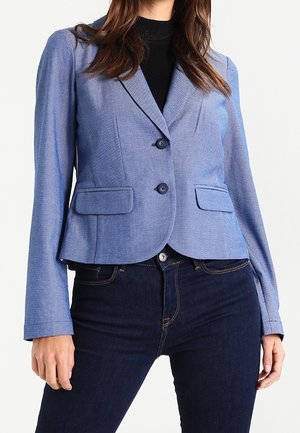 Kvinde iført en tætsiddende, lyseblå blazer med to knapper, lommer med klap og mørkeblå jeans, stående mod en ensfarvet baggrund.