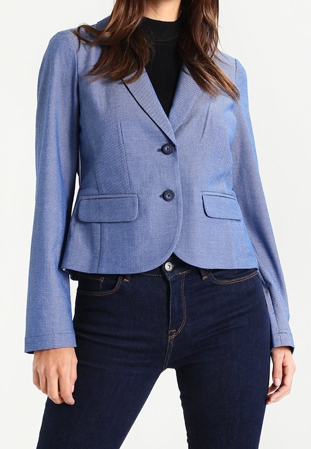 Femme portant un blazer ajusté bleu clair avec deux boutons, poches à rabat et jean bleu foncé, debout devant un fond uni.