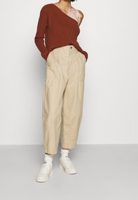 Rust-colored ribbed sweater with one shoulder exposed, paired with beige high-waisted cargo pants and white sneakers with textured soles.