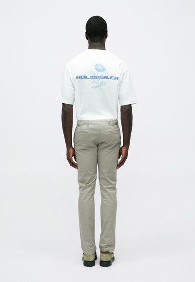 Λευκό κοντομάνικο μπλουζάκι με μπλε γραφικό κείμενο και σχέδιο φυτού στο πίσω μέρος, συνδυασμένο με ανοιχτό μπεζ παντελόνι και πράσινα παπούτσια.