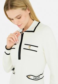Maglione lavorato a maglia in crema con dettagli neri. Presenta un colletto con zip, due tasche sul petto con bottoni e un logo ricamato sull'orlo.