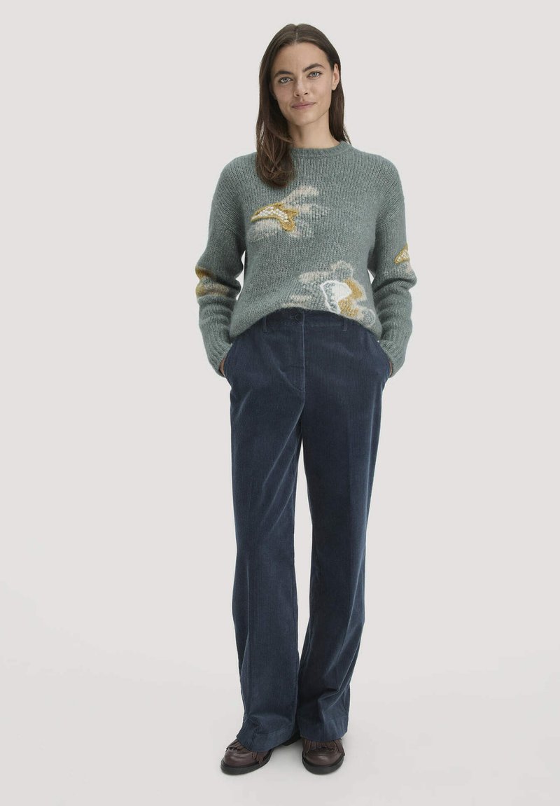 Vrouw staat met haar handen in de zakken van een marineblauwe corduroy broek, gekleed in een grijze trui met witte en gele bloemenpatronen en bruine schoenen.