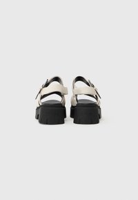 Footwear: back view of sandals with beige straps, black chunky sole, adjustable buckles, and textured upper for grip.