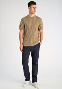 Μπεζ πλεκτό πουλόβερ με κοντά μανίκια και υφασμένο μοτίβο καλώδιου, σε συνδυασμό με ναυτικό μπλε chino παντελόνια και λευκά αθλητικά παπούτσια. Απλός και λειτουργικός σχεδιασμός.