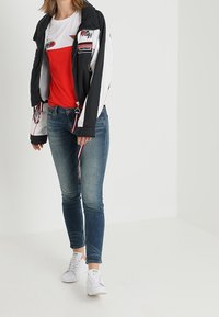 Chaleco cortavientos en blanco y negro con detalles de parches, camiseta roja, jeans azules y zapatillas blancas, con un diseño ajustado y casual.