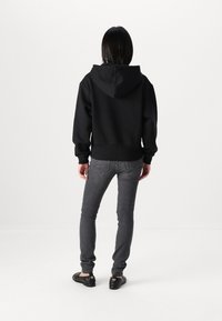 Felpa nera con cappuccio, dal taglio comodo, con polsini e orlo a coste. Indossata con jeans grigi scuri e scarpe nere. Vista da dietro.