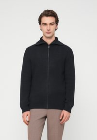 Στενόπλεκτο πουλόβερ ζακέτα σε σκούρο ναυτικό μπλε με ψηλό γιακά, χαρακτηριστική ριμπ υφή και δομημένες ραφές. Λεπτή γραμμή, μακριά μανίκια και χωρίς εμφανές λογότυπο.