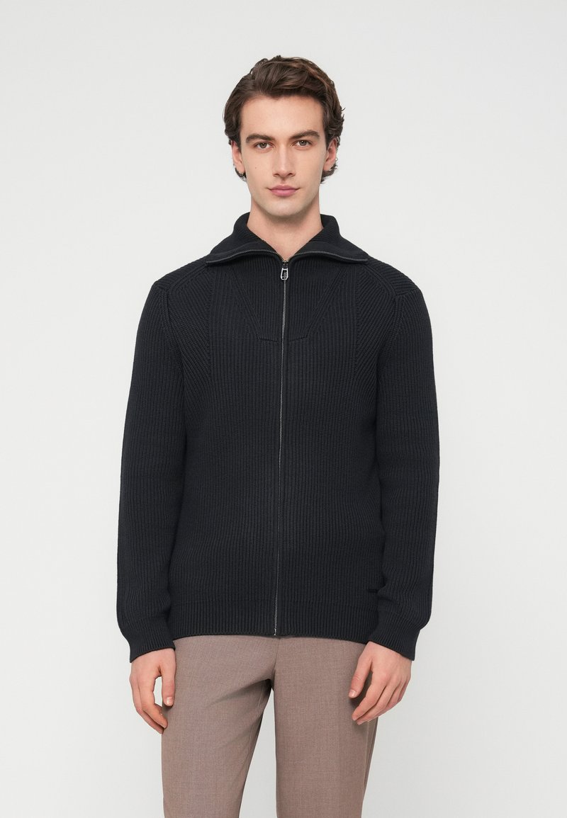 Στενόπλεκτο πουλόβερ ζακέτα σε σκούρο ναυτικό μπλε με ψηλό γιακά, χαρακτηριστική ριμπ υφή και δομημένες ραφές. Λεπτή γραμμή, μακριά μανίκια και χωρίς εμφανές λογότυπο.