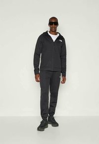 Felpa nera con zip e pantaloni jogger abbinati realizzati in un tessuto morbido ed elastico, con dettagli del logo e una silhouette aderente. Sneakers nere completano il look.