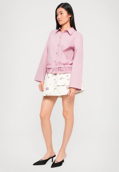 Różowa bluzka z długim rękawem, z falbaną u dołu, zapinana na guziki, zestawiona z białą spódnicą w kwiaty i czarnymi szpilkami z wąskim noskiem.