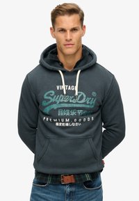 Donkerblauwe hoodie met trekkoorden, met een voorzak en grafische tekst in groen en wit. Gedragen over een geruit shirt.