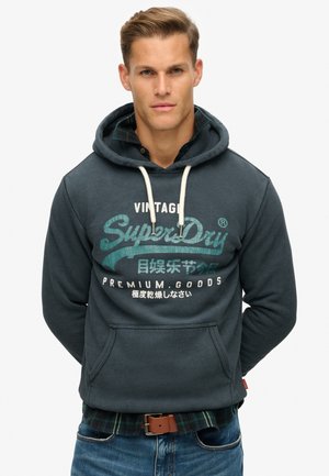 Donkerblauwe hoodie met trekkoorden, met een voorzak en grafische tekst in groen en wit. Gedragen over een geruit shirt.