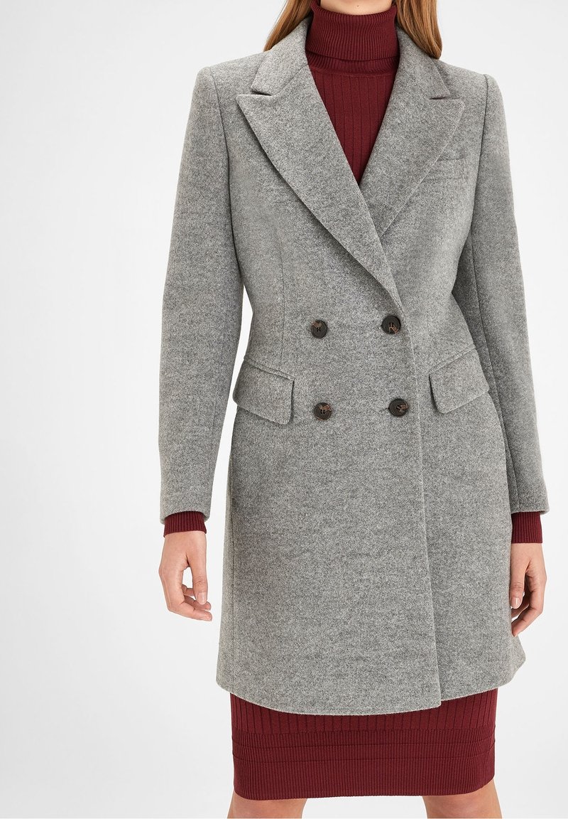 Manteau en laine mélangée gris avec un design croisé, des revers en pointe, deux poches à l'avant et des boutons noirs, porté sur une robe côtelée bordeaux.