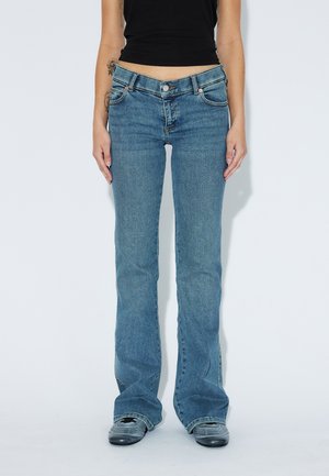 Osoba nosząca niebieskie jeansy typu bootcut i czarne trampki, stojąca na jednolitym białym tle z rękami swobodnie opuszczonymi wzdłuż boków.