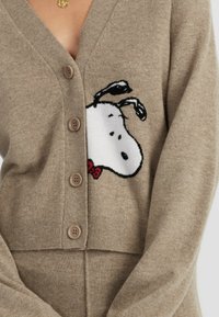 Cardigan beige con scollo a V, caratterizzato da un ricamo di Snoopy sul lato sinistro, cinque bottoni marroni e polsini a costine sulle maniche.