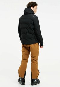 Schwarze Pufferjacke mit Kapuze, die eine glatte Textur und einen lässigen Schnitt aufweist. Dazu braune Skihosen und schwarze Skischuhe.