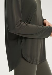 Maglietta sportiva a maniche lunghe in tessuto grigio scuro, con orlo curvato e texture liscia. Mostrata parzialmente insieme a leggings abbinati.
