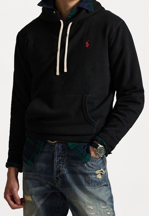 Schwarzer Hoodie mit einer Fronttasche, cremefarbenen Kordeln und rotem Logo. Kombiniert mit einem grünen karierte Hemd und ausgewaschenen blauen Jeans.