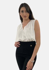 Blusa bianca senza maniche con scollatura a V, orlo arricciato; abbinata a pantaloni neri a vita alta con un accento di bottone dorato. Tessuto dalla texture liscia.