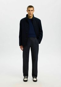 Cappotto di lana blu navy con colletto alto e quattro bottoni, abbinato a un maglione blu scuro e pantaloni scuri a trama. Scarpe nere e argento.