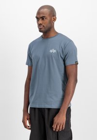 Camiseta de algodón azul con mangas cortas, cuello redondo y un pequeño diseño gráfico blanco en el pecho, combinada con pantalones cargo negros.