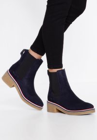 Botas de tobillo de ante azul marino con paneles laterales elásticos, un tacón cuadrado y una suela texturizada. Un ribete rojo y blanco a lo largo de la suela añade detalle.