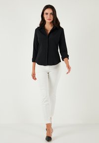 Black button-up shirt with long sleeves, paired with white fitted pants; model wearing black pointed-toe heels and layered gold necklaces.
