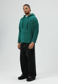 Sweat à capuche en molleton vert sarcelle avec cordons, poche frontale, porté avec un jean noir à jambes larges et des chaussures noires avec des lacets roses.