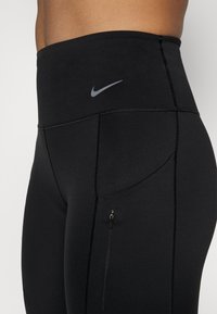 Leggings negros de cintura alta con una textura suave, que cuentan con un pequeño bolsillo con cremallera y un logo de Nike en gris en la cintura.